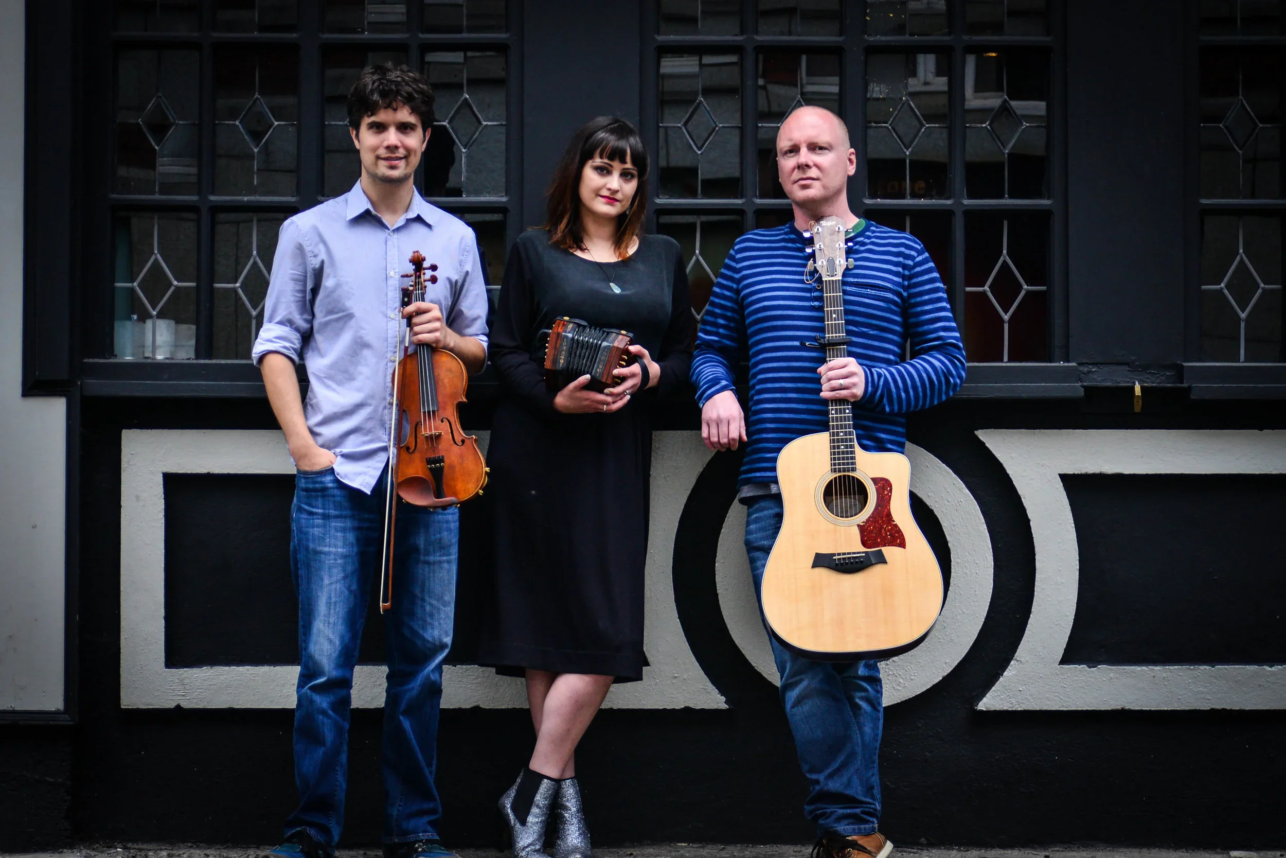 Nathan, Mairéad, and John standing with fiddle, concertina, and guitar outside a dark-painted building with stained glass windows
